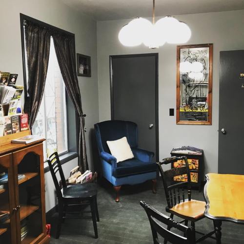 Cozy room with a blue armchair, wooden table, black chairs, and a bookshelf, featuring gray walls and warm lighting.