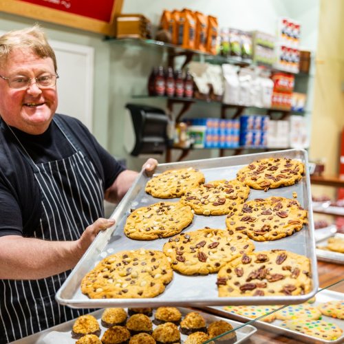 A smiling baker holds a tray of large cookies in a cozy bakery, with various baked goods displayed in the background.