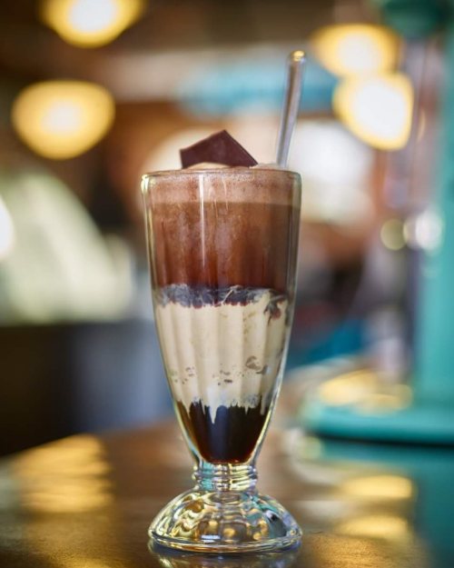 A tall glass filled with layers of chocolate syrup, ice cream, and topped with a chocolate square, sitting on a wooden table.