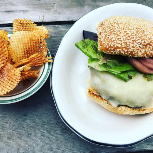 A sesame seed bun burger with lettuce, cheese, and ham, served with a side of wavy potato chips.
