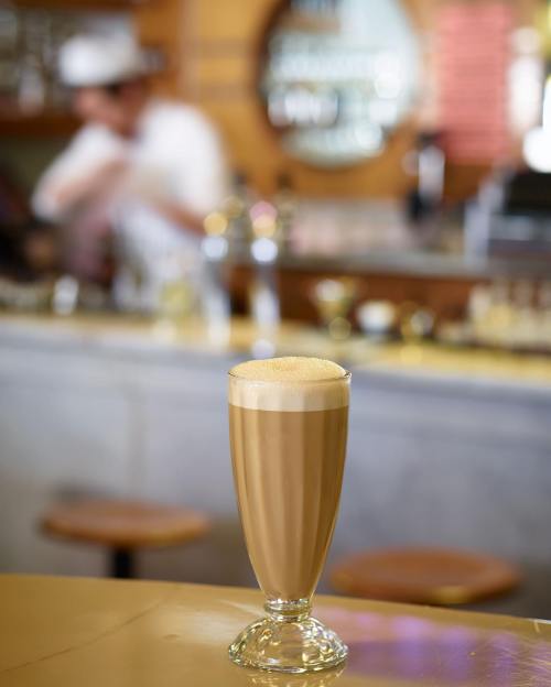 A tall glass of frothy coffee sits on a counter, with a blurred barista in the background.