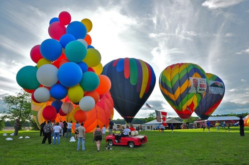 Colorful hot air balloons inflate against a cloudy sky, with a crowd and a small red car in a grassy field.