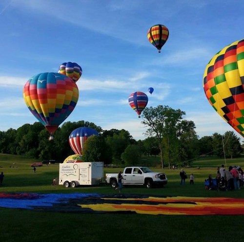 Colorful hot air balloons ascend in a clear sky, with a truck and people on the ground in a park setting.