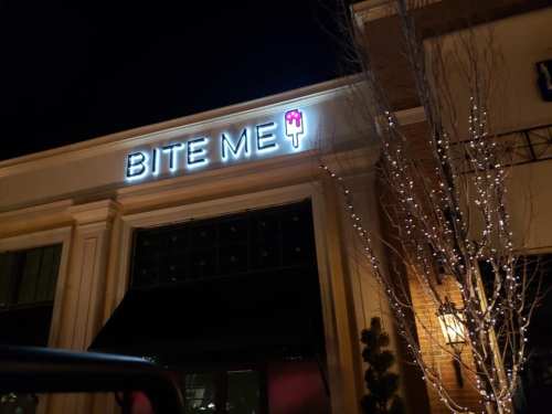 Neon sign reading "BITE ME" with a popsicle icon, illuminated at night next to a building decorated with lights.