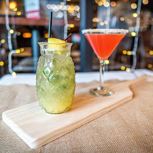 Two colorful cocktails on a wooden serving board, with festive lights in the background.