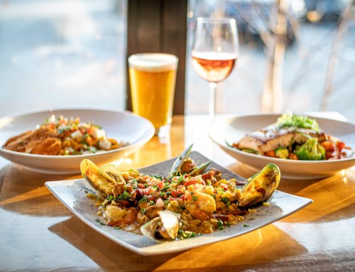 A table setting with a seafood dish, a plate of salmon, a beer, and a glass of rosé wine, all in natural light.