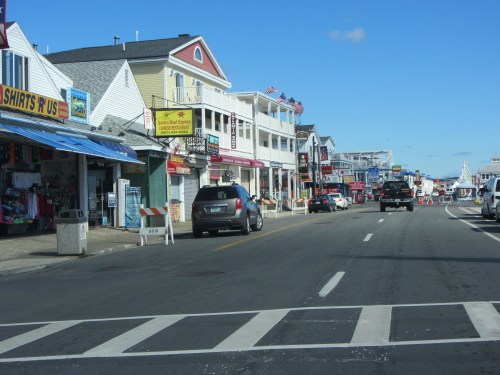 A seaside street lined with shops and colorful buildings, with a clear blue sky and a few parked cars.