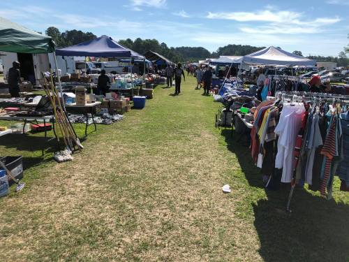 A bustling outdoor market with tents, tables, and clothing racks, filled with shoppers and various goods.