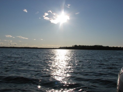 A bright sun reflects on calm water, with a distant shoreline under a clear blue sky and scattered clouds.
