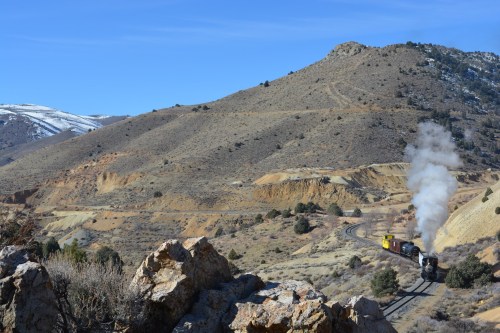 A steam train travels along a winding track through a mountainous landscape with rocky terrain and sparse vegetation.
