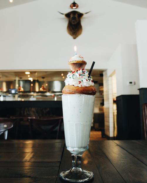 A tall milkshake topped with a cupcake and sprinkles, with a candle, set against a rustic restaurant backdrop.
