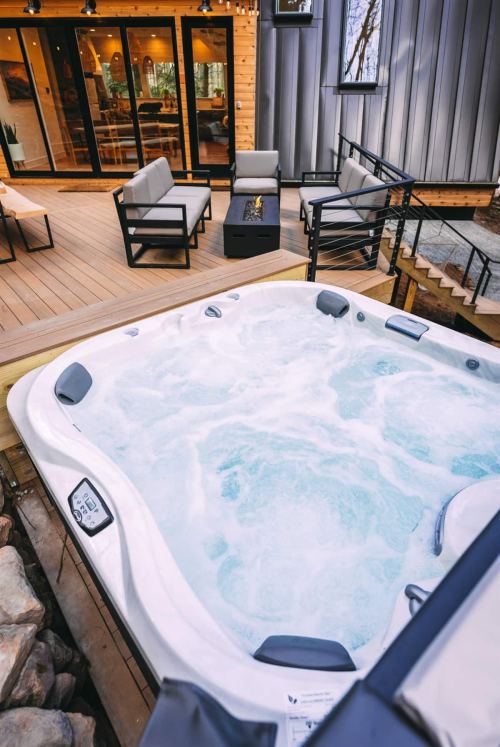 A hot tub with bubbling water on a wooden deck, surrounded by modern outdoor seating and a fire pit.