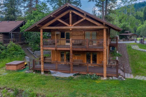 A rustic wooden cabin with two levels, surrounded by trees and a grassy area, featuring a hammock and hot tub.
