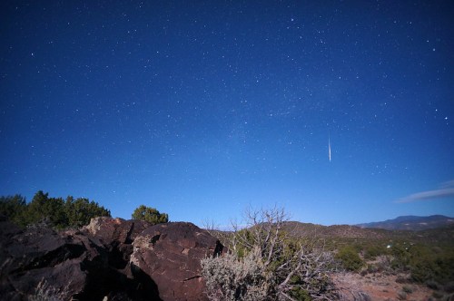 A clear night sky filled with stars, featuring a shooting star over rocky terrain and distant mountains.