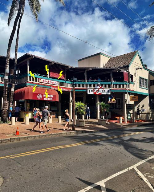 A street view of shops with colorful awnings, palm trees, and people walking by under a partly cloudy sky.