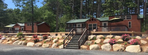 Three rustic cabins with green roofs, surrounded by trees and colorful flower beds, set on a gravel path.