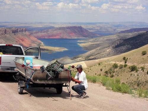 A person prepares a boat on a trailer near a scenic overlook of a lake and mountains under a cloudy sky.