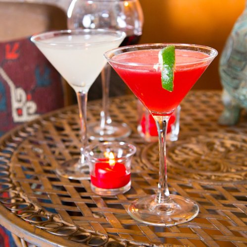 Two colorful cocktails on a woven table, with a lit candle nearby, set in a cozy atmosphere.