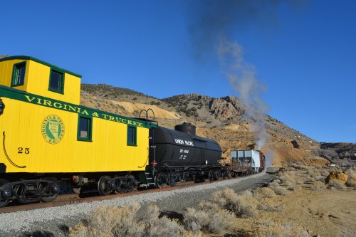 A vintage yellow train caboose and black tanker car on a track, with smoke rising in a mountainous desert landscape.