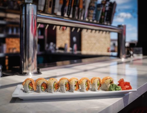 A plate of sushi rolls with orange topping, served with wasabi and ginger, on a marble countertop near a bar.
