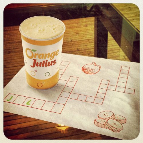 A cup of Orange Julius on a table next to a crossword puzzle sketch with an orange and cucumber drawings.