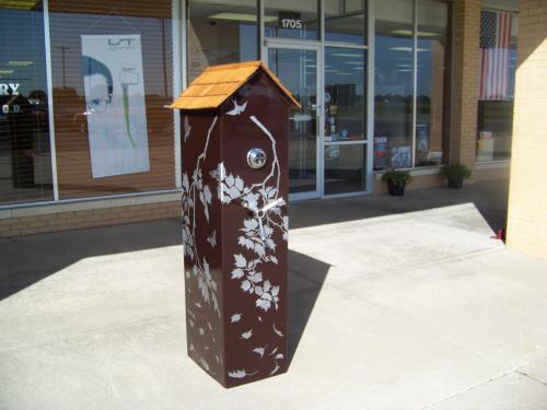 A decorative brown mailbox with a wooden roof and leaf designs, placed outside a storefront.