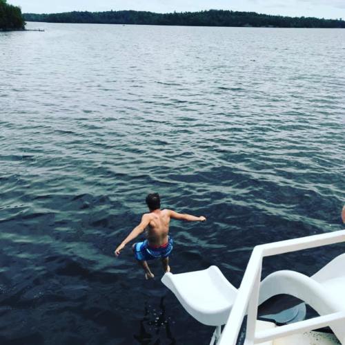 A person jumps off a water slide into a lake, surrounded by trees and a cloudy sky.