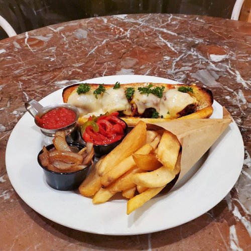 A plate of cheesy sausage on a toasted bun, served with fries, grilled onions, and a side of salsa.