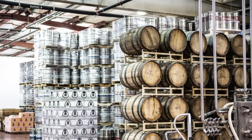 A warehouse filled with stacked barrels, kegs, and pallets of canned goods, showcasing a brewing or distilling facility.