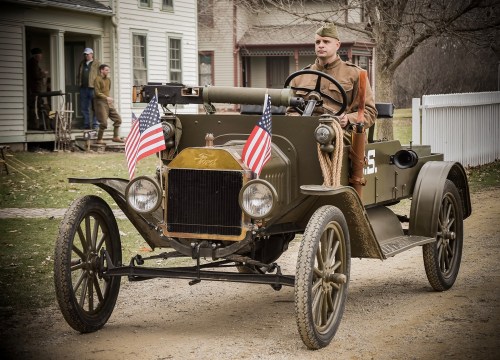 A soldier drives a vintage military vehicle adorned with American flags on a dirt road near historic buildings.