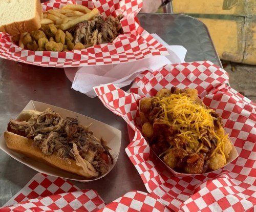 A table with red-checkered paper featuring pulled pork sandwiches, tater tots with cheese, and fried okra.