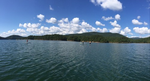 A serene lake scene with paddleboarders on calm water, surrounded by green hills and a blue sky with fluffy clouds.