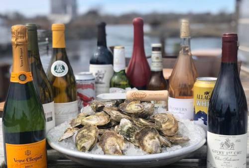 A platter of oysters on ice surrounded by various bottles of wine and beer, set against a scenic outdoor backdrop.