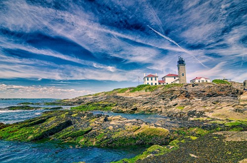A scenic lighthouse on rocky shores under a vibrant blue sky with wispy clouds and green moss on the rocks.