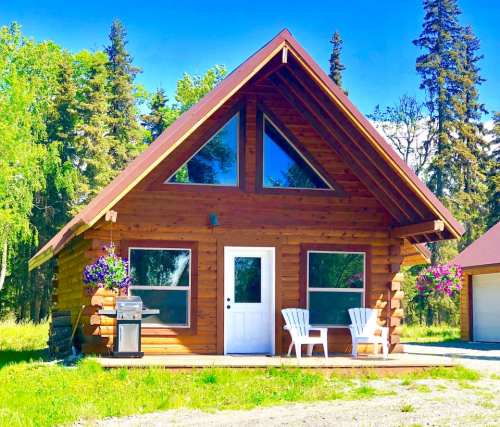 A cozy wooden cabin with large windows, surrounded by trees, featuring a grill and two white chairs on the porch.