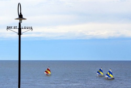 A signpost for Bay St. Louis stands by the water, with colorful sailboats sailing in the distance under a cloudy sky.