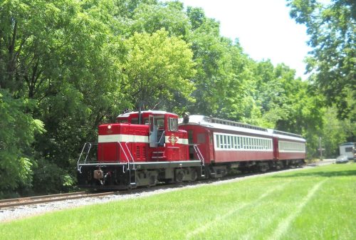 A red and white train engine pulls a matching passenger car along a green, tree-lined track on a sunny day.