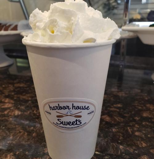 A white cup filled with whipped cream, featuring a logo for Harbor House Sweets on a dark countertop.