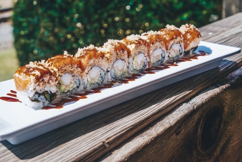 A long platter of sushi rolls drizzled with sauce, garnished with crispy toppings, set on a wooden table.