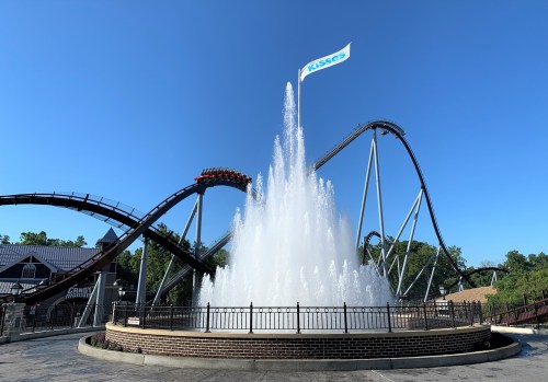 A roller coaster loops above a large water fountain, with a blue sky in the background and a flag that reads "KISSES."