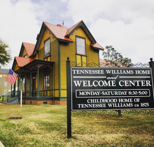 Historic yellow and green house with a sign for the Tennessee Williams Home and Welcome Center, featuring an American flag.
