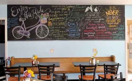 Chalkboard mural with inspirational quotes and a bicycle illustration, above empty tables with flowers.