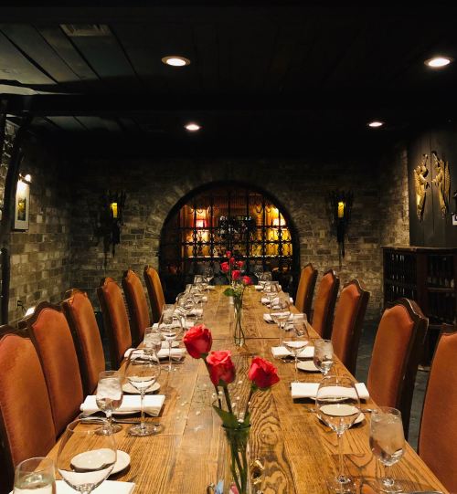 A long wooden dining table set with glasses and napkins, adorned with red roses, in a dimly lit, rustic restaurant.
