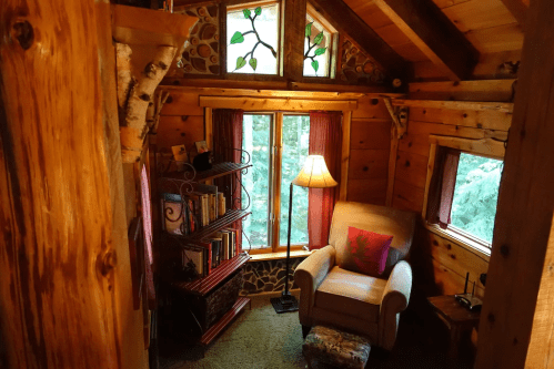 Cozy wooden interior of a cabin with a reading nook, bookshelf, lamp, and large windows overlooking greenery.