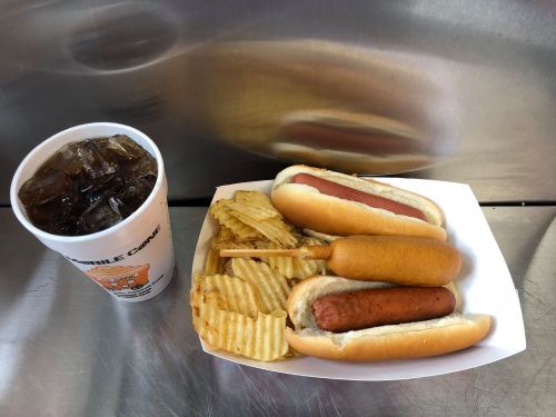 A tray with two hot dogs, a corn dog, crinkle-cut chips, and a cup of iced soda on a metal surface.