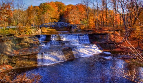 A serene waterfall cascades over rocks, surrounded by vibrant autumn foliage and a blue bridge in the background.