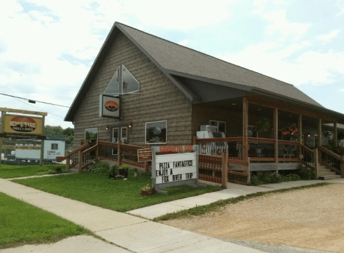 A rustic restaurant with a wooden deck and sign advertising pizza, surrounded by greenery and a blue sky.