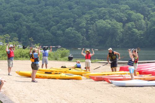 Group of people in life jackets preparing kayaks on a sandy beach by a lake, surrounded by green hills.