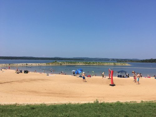 A sunny beach scene with people swimming and relaxing, umbrellas, and a calm lake in the background.