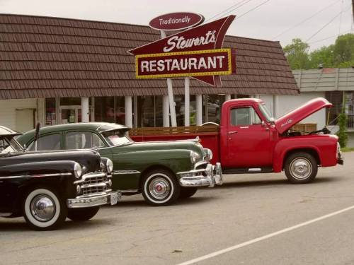 Classic cars parked in front of Stewart's Restaurant, featuring a black sedan, green car, and a red pickup truck.